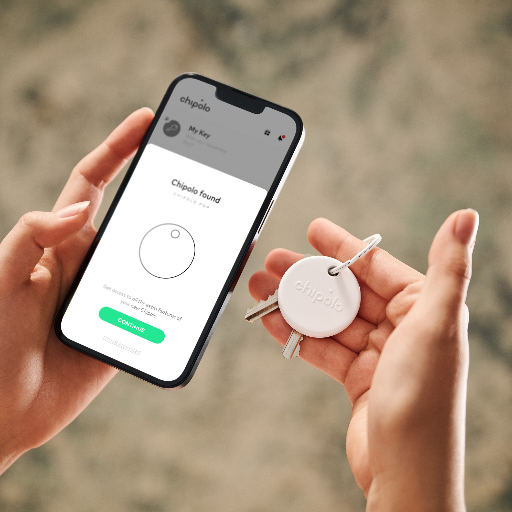 A person holds a smartphone showing a Chipolo app setup screen and a set of keys with a white Chipolo tracker.