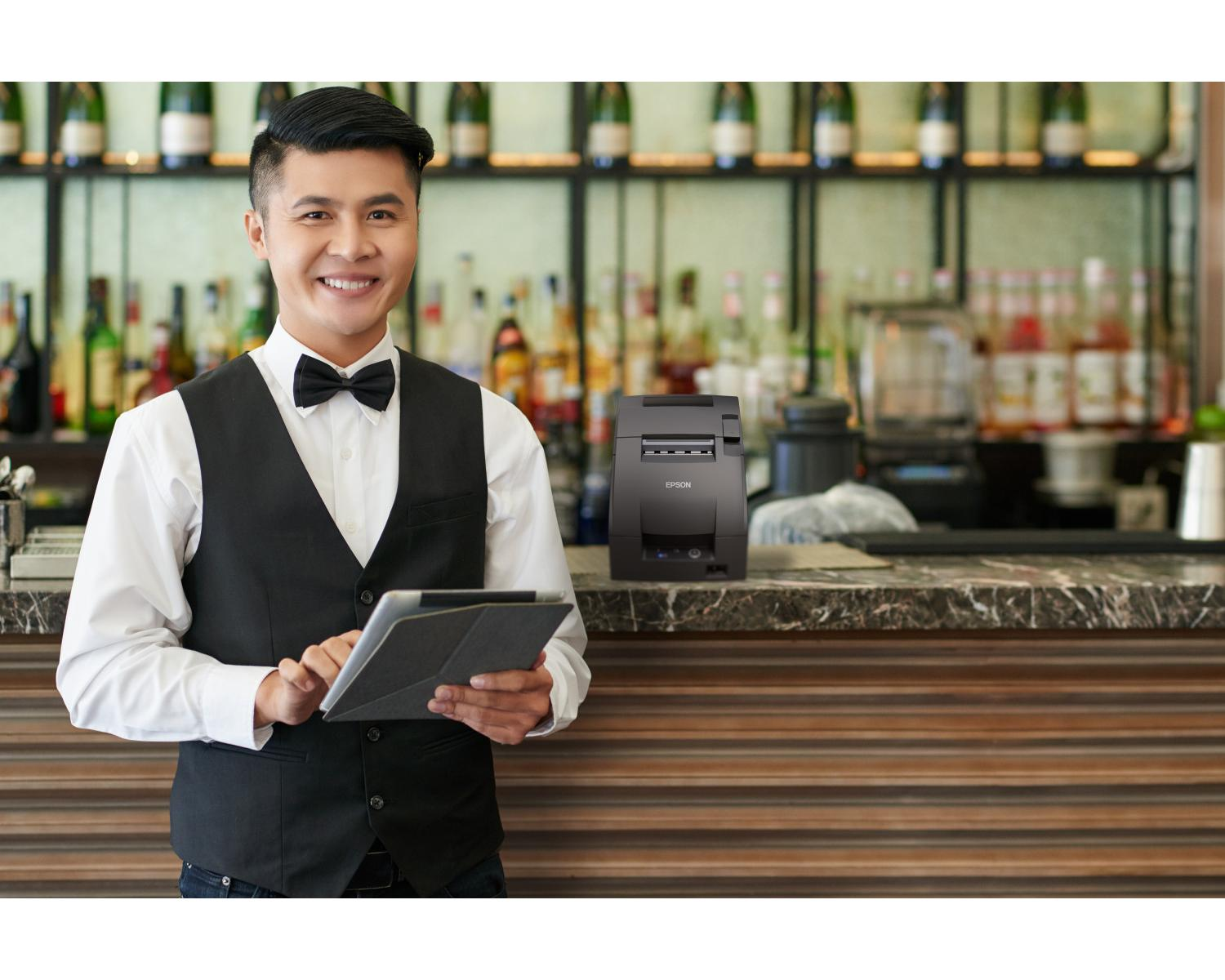 A smiling waiter in a vest and bow tie holds a tablet in a bar with a receipt printer on the counter.