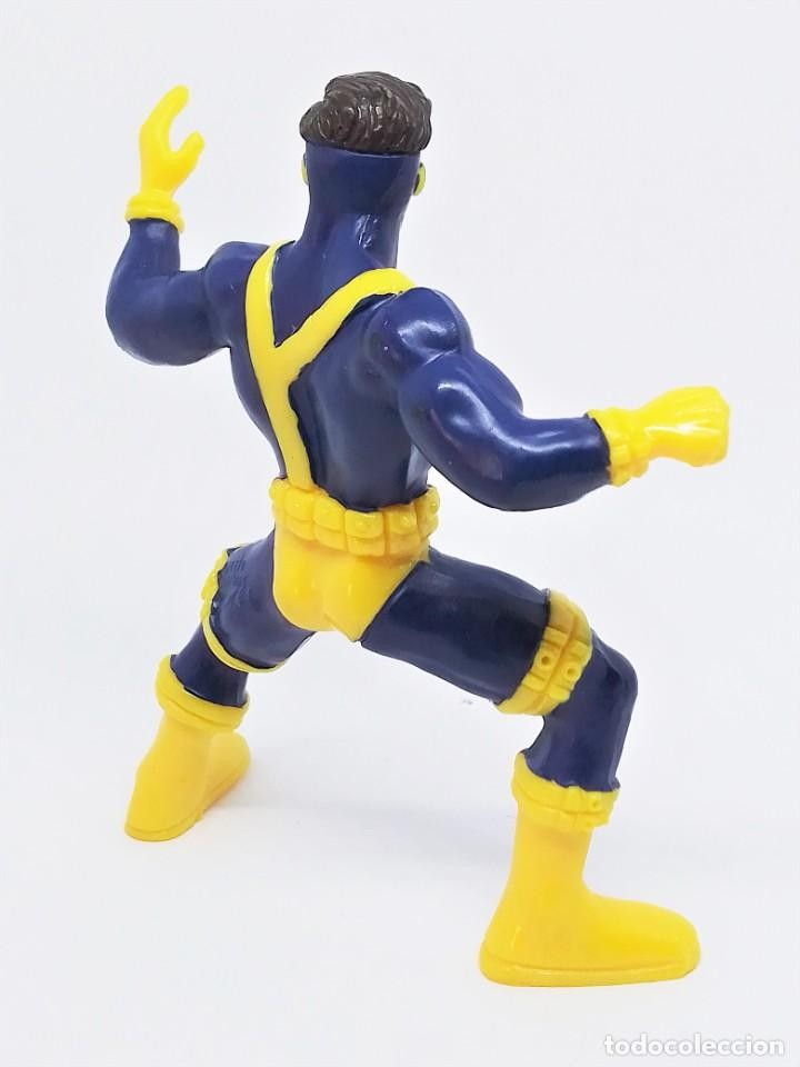 Vista trasera de una figura de acción de Cíclope con traje azul y amarillo, en postura de acción sobre fondo blanco.