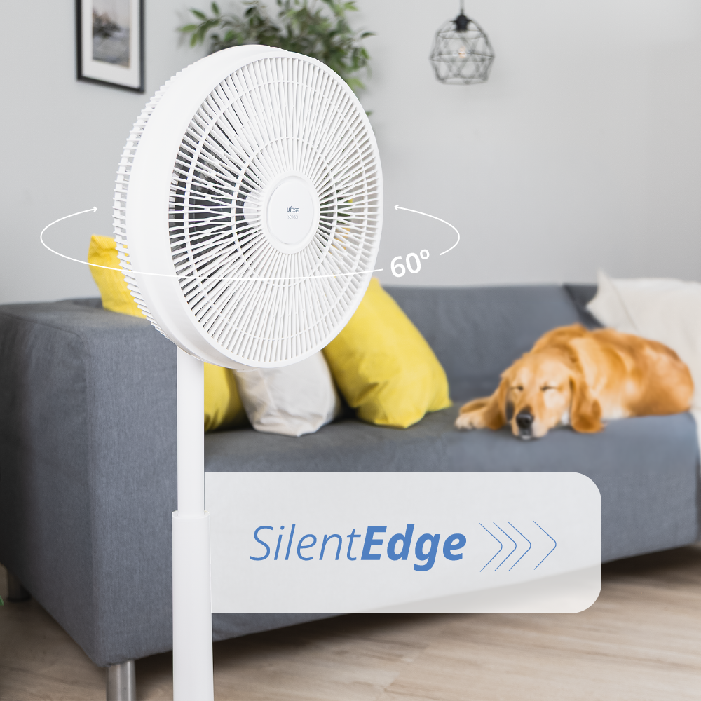 Un ventilador de pie blanco con oscilación de 60 grados en una sala con un perro durmiendo en un sofá al fondo.