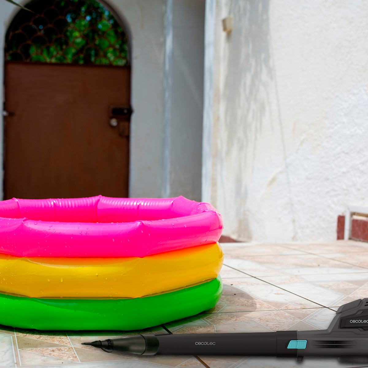 Una piscina hinchable de colores está en un patio de azulejos junto a un dispositivo negro.