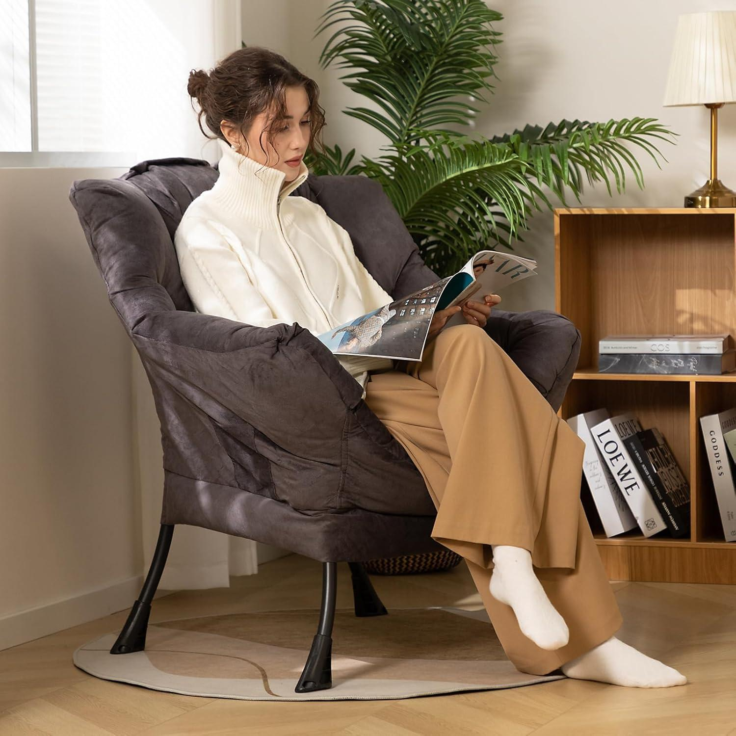 Una donna con maglione bianco e pantaloni beige siede in una poltrona grigia leggendo una rivista.