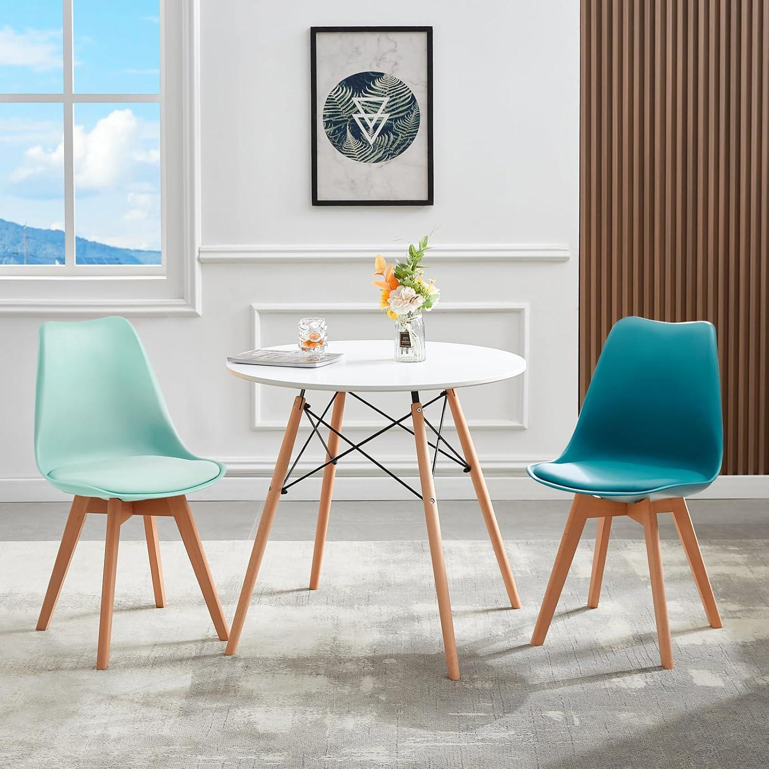 Un tavolo rotondo bianco con gambe in legno è posizionato tra una sedia verde menta e una blu su un tappeto grigio.