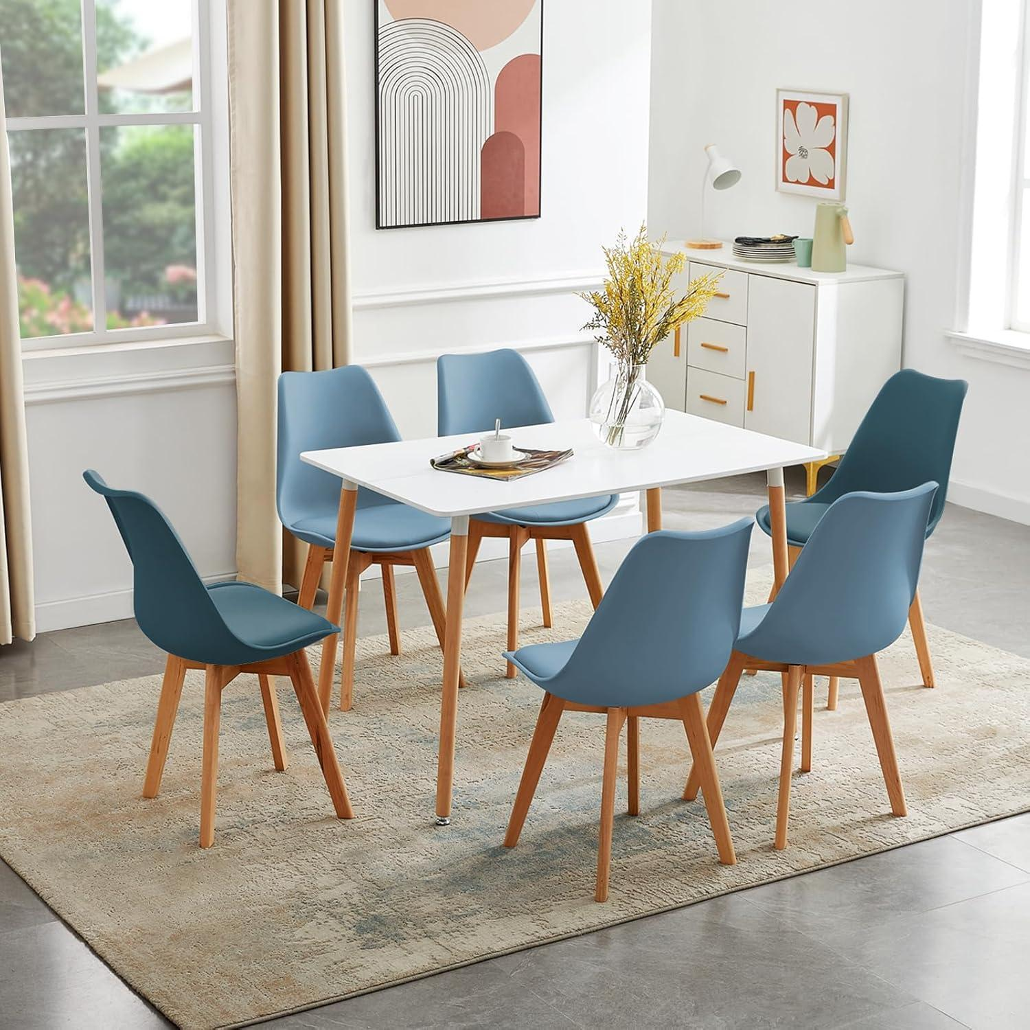 Un tavolo da pranzo bianco con sei sedie blu su un tappeto fantasia in una stanza luminosa.