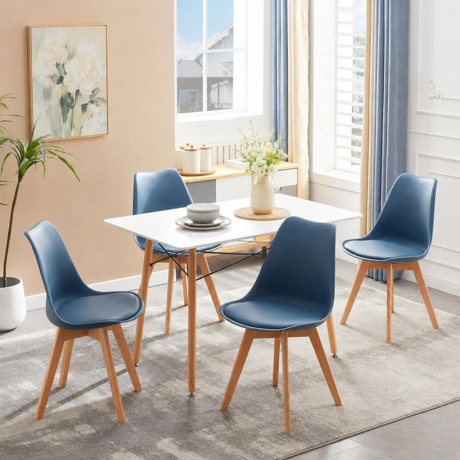 Luminosa zona pranzo con tavolo bianco, quattro sedie blu, una pianta e un quadro incorniciato.