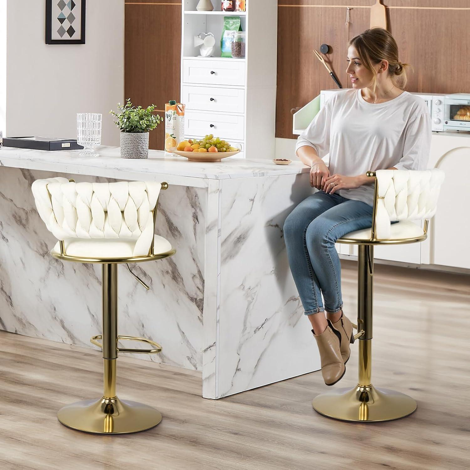Una donna siede su uno sgabello da bar bianco con struttura dorata a un bancone di marmo in una cucina moderna.
