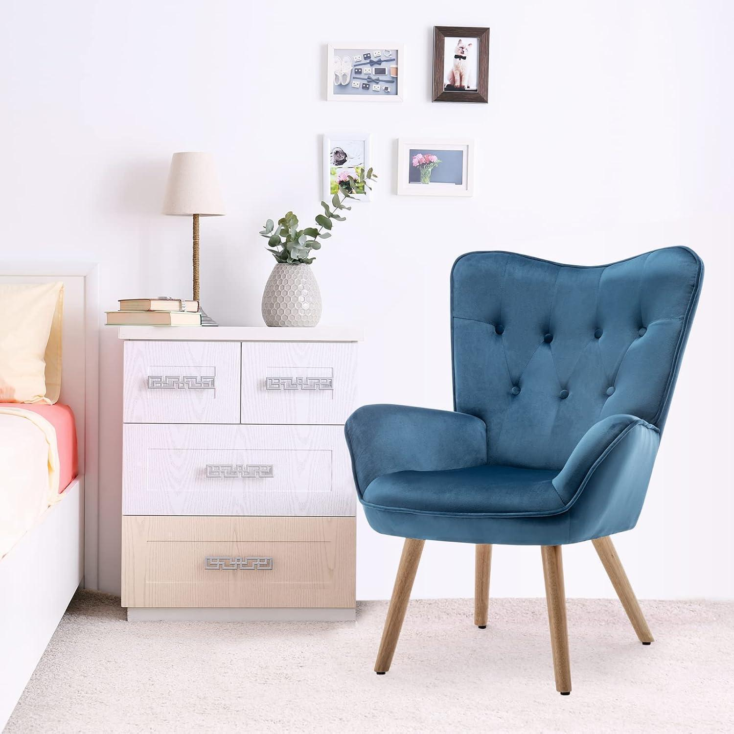 Una poltrona in velluto blu con gambe in legno si trova in una camera luminosa accanto a un comò bianco.