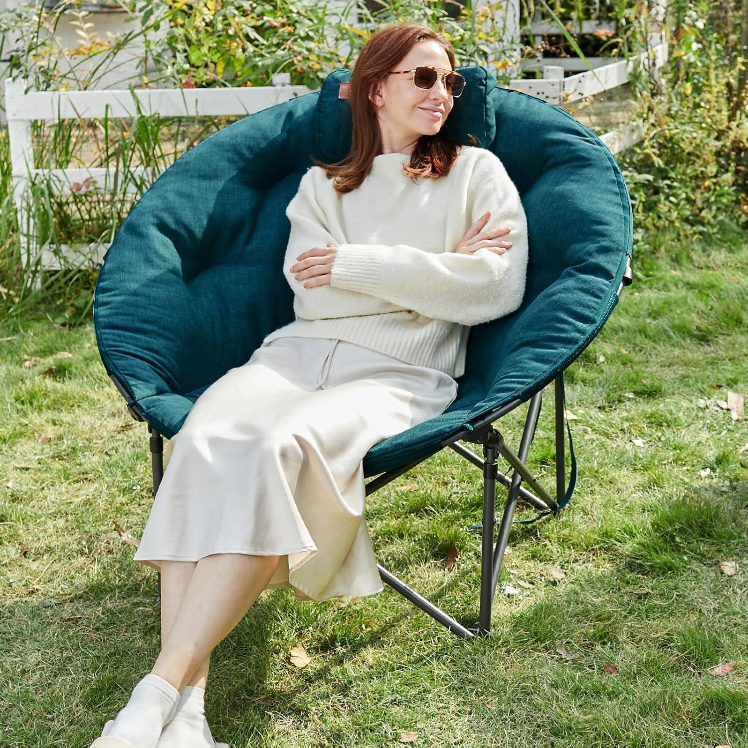 Una donna con maglione bianco e gonna siede comodamente in una sedia pieghevole verde scuro sul prato.