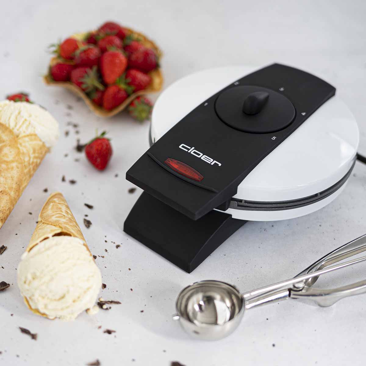 A white and black ice cream cone maker sits on a table with ice cream cones, strawberries, and an ice cream scoop.