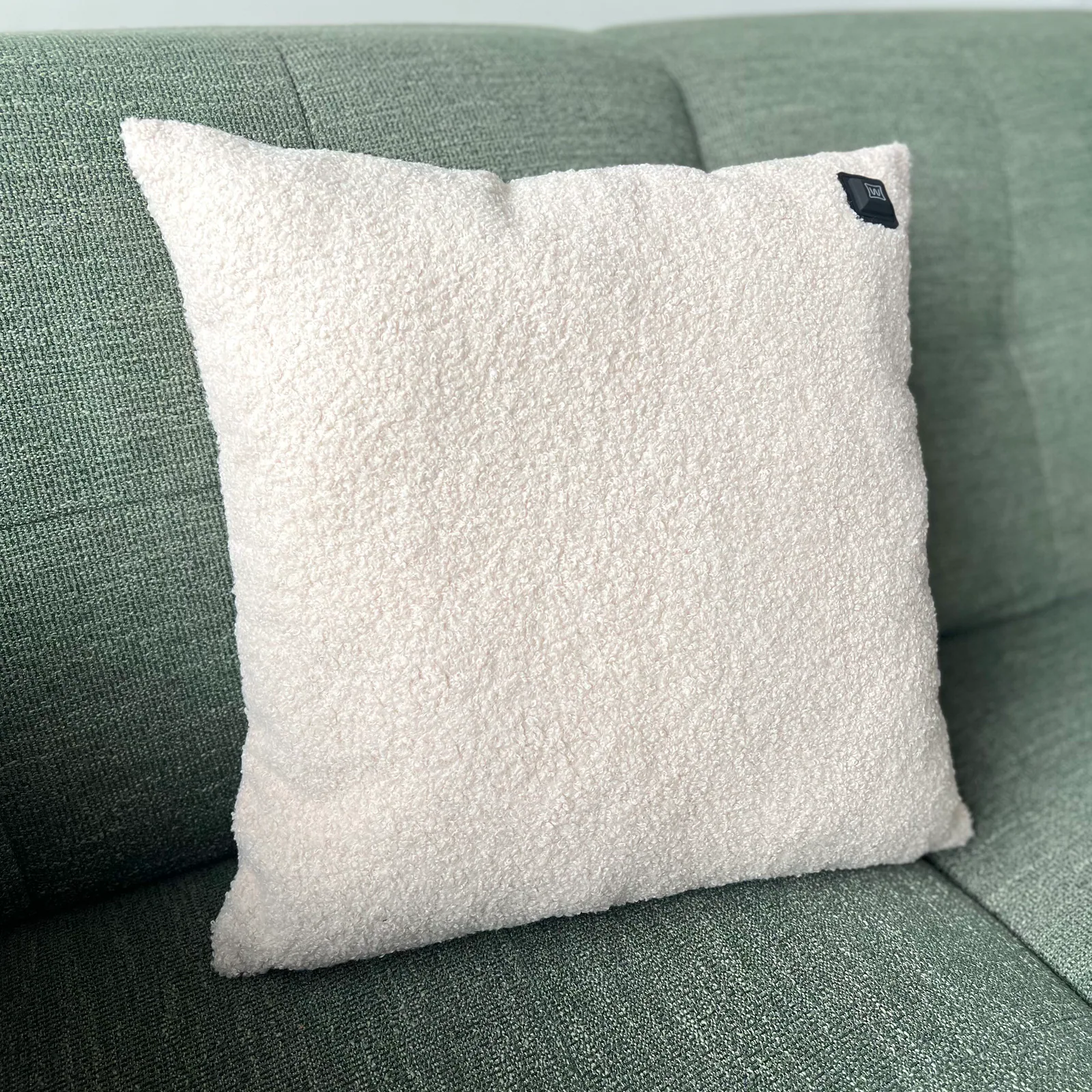 Ein flauschiges weißes Kissen mit einem kleinen schwarzen Etikett liegt auf einem grünen Sofa.