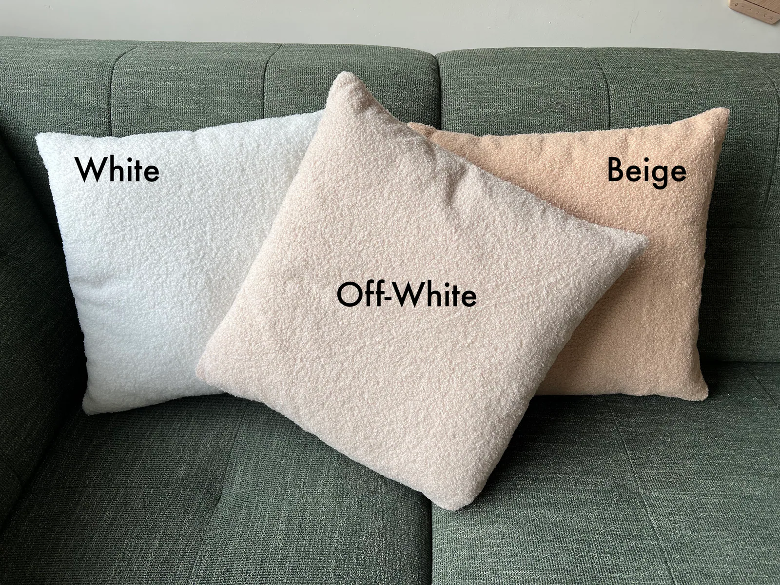 Drei flauschige Kissen in Weiß, gebrochenem Weiß und Beige liegen auf einem grünen Sofa.