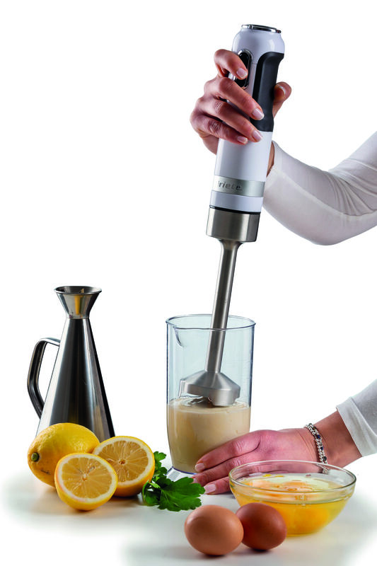Een hand bedient een staafmixer in een maatsbeker, omringd door citroenen, eieren en een metalen kan.