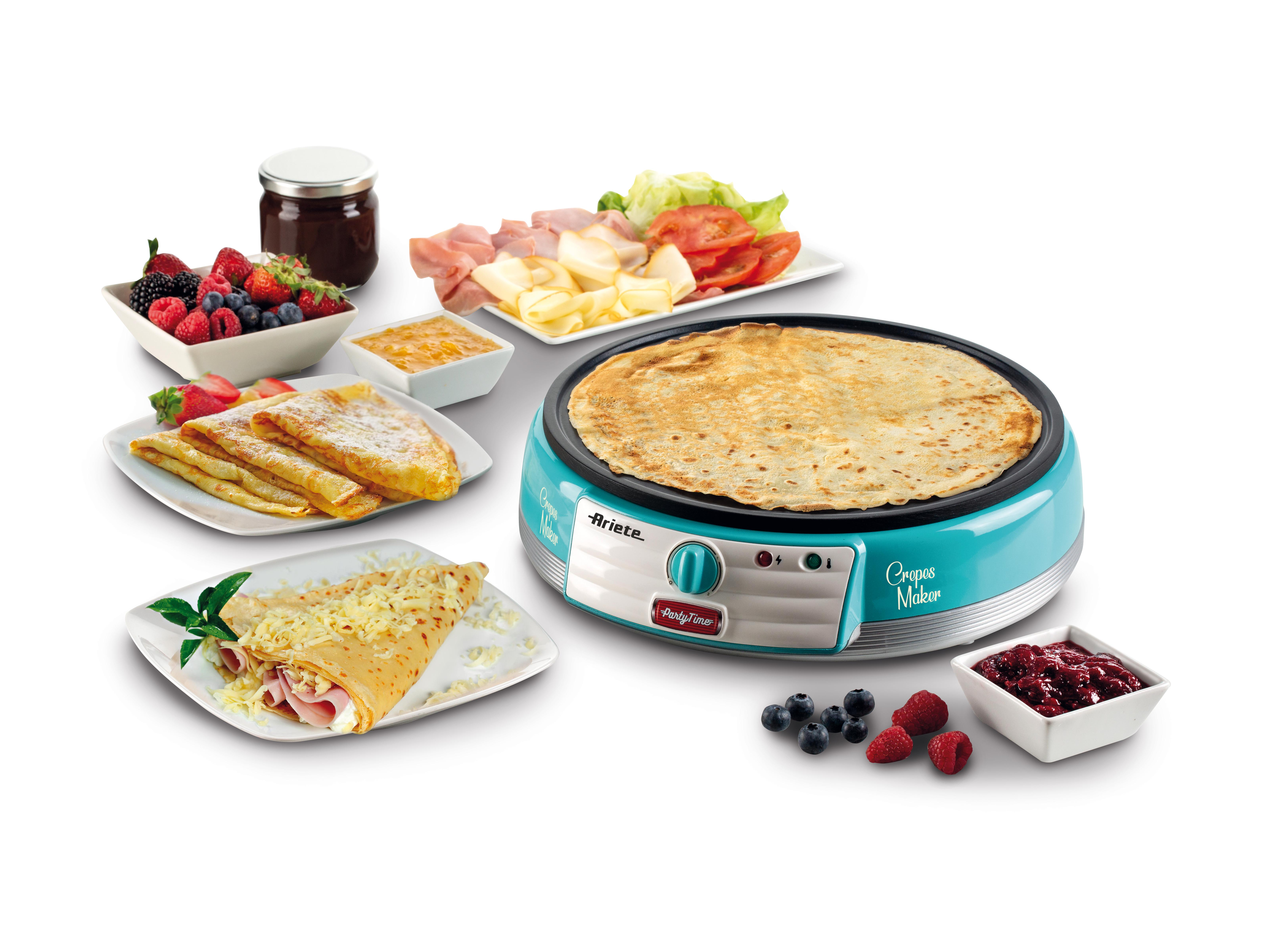 A blue crepe maker on a table, surrounded by filled crepes, fresh fruit, ham, and cheese.