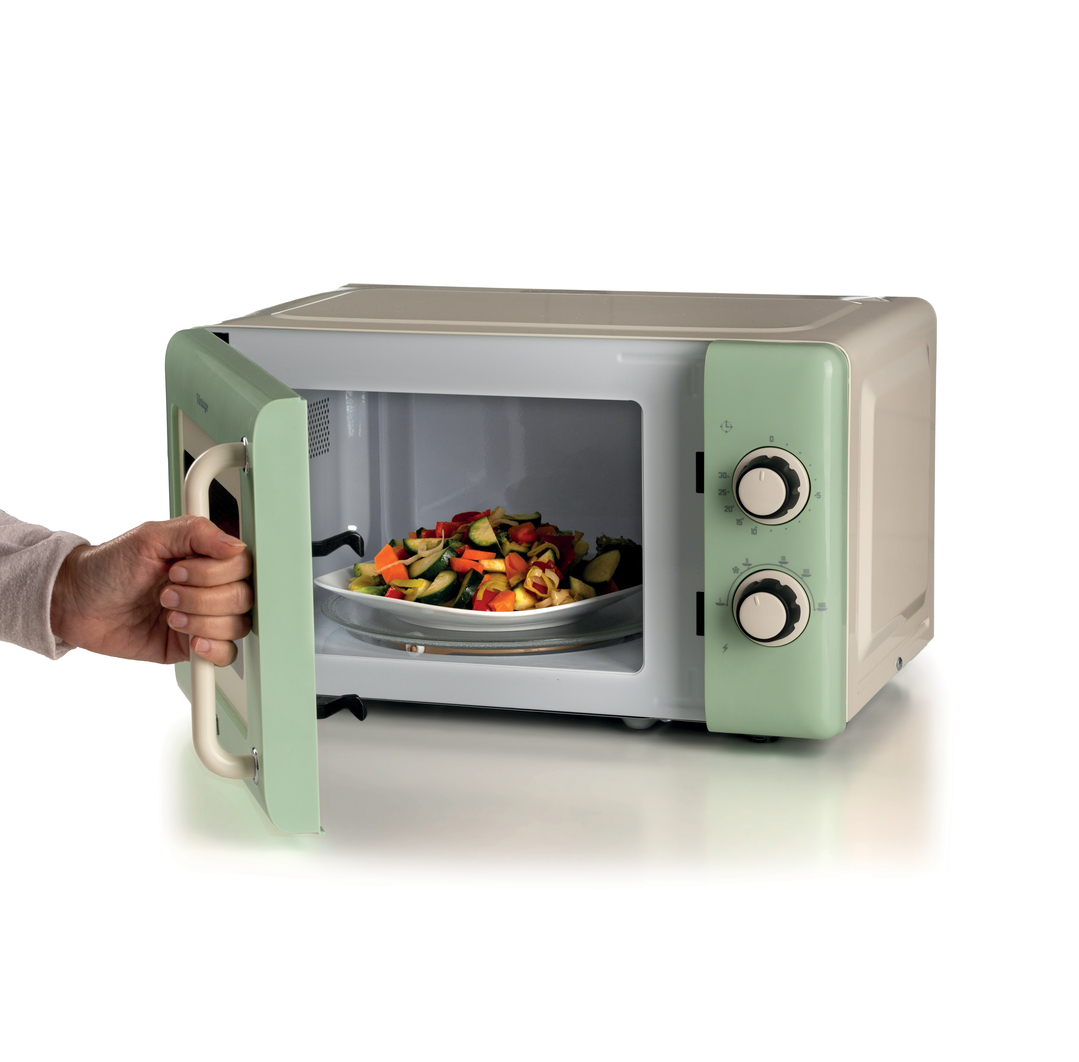 Una mano abre la puerta de un microondas vintage verde claro con un bol de verduras dentro.