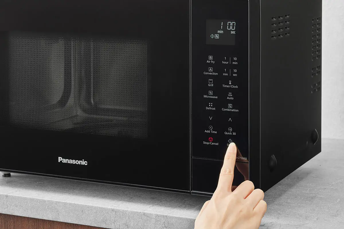 Ręka naciska przycisk start na czarnej mikrofalówce Panasonic ustawionej na szarym blacie.