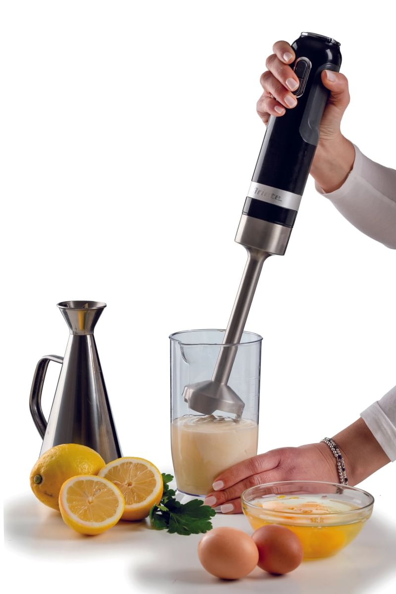 Een hand bedient een zwarte staafmixer boven een bekers met vloeistof, naast citroenen, eieren en een kan.