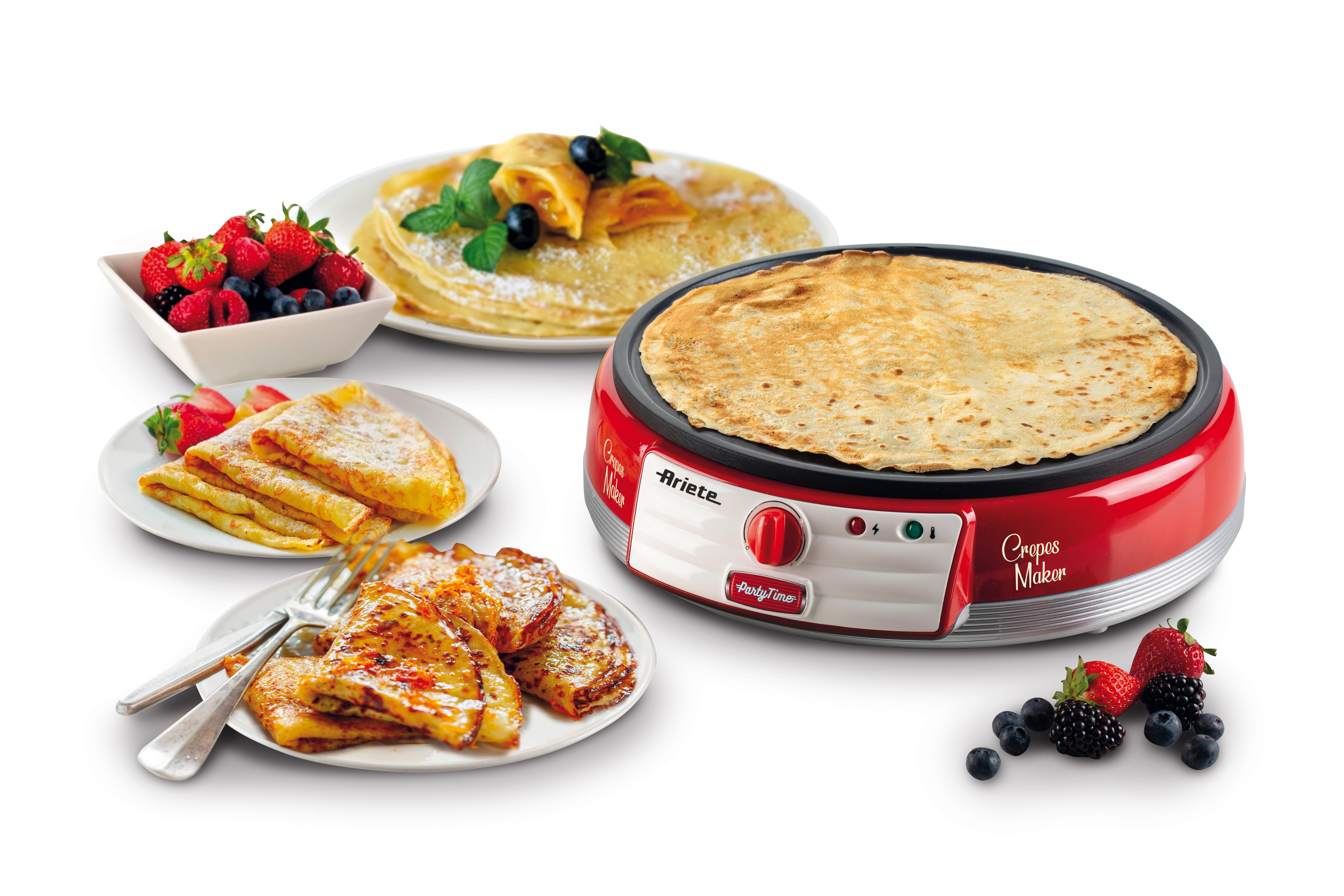 A red crepe maker sits beside plates of fresh crepes, berries, and cutlery on a white background.