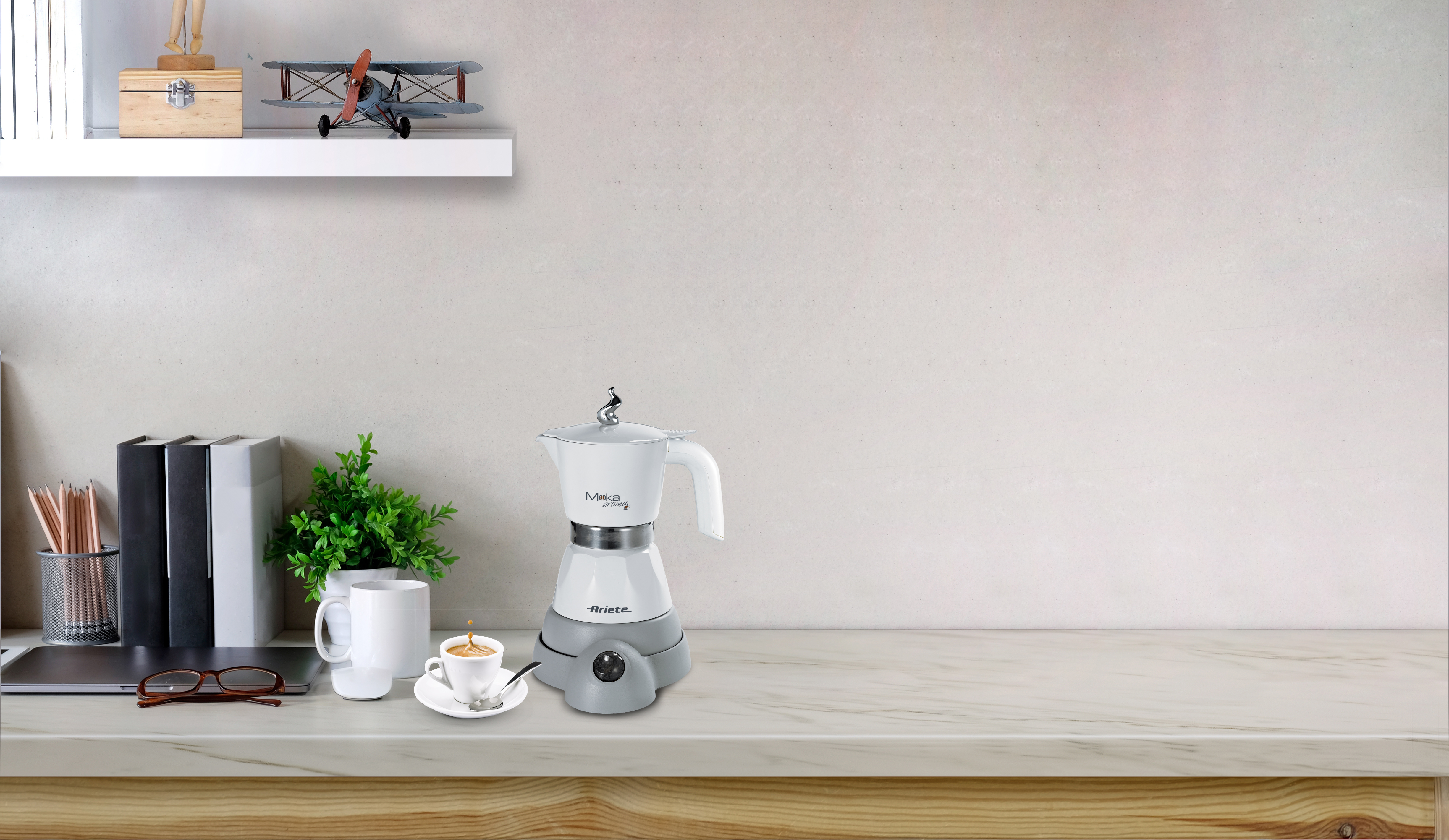 Eine weiße Moka-Kaffeemaschine steht auf einer Marmorarbeitsplatte neben einer Tasse Kaffee, Büchern und Pflanzen vor einer schlichten Wand.
