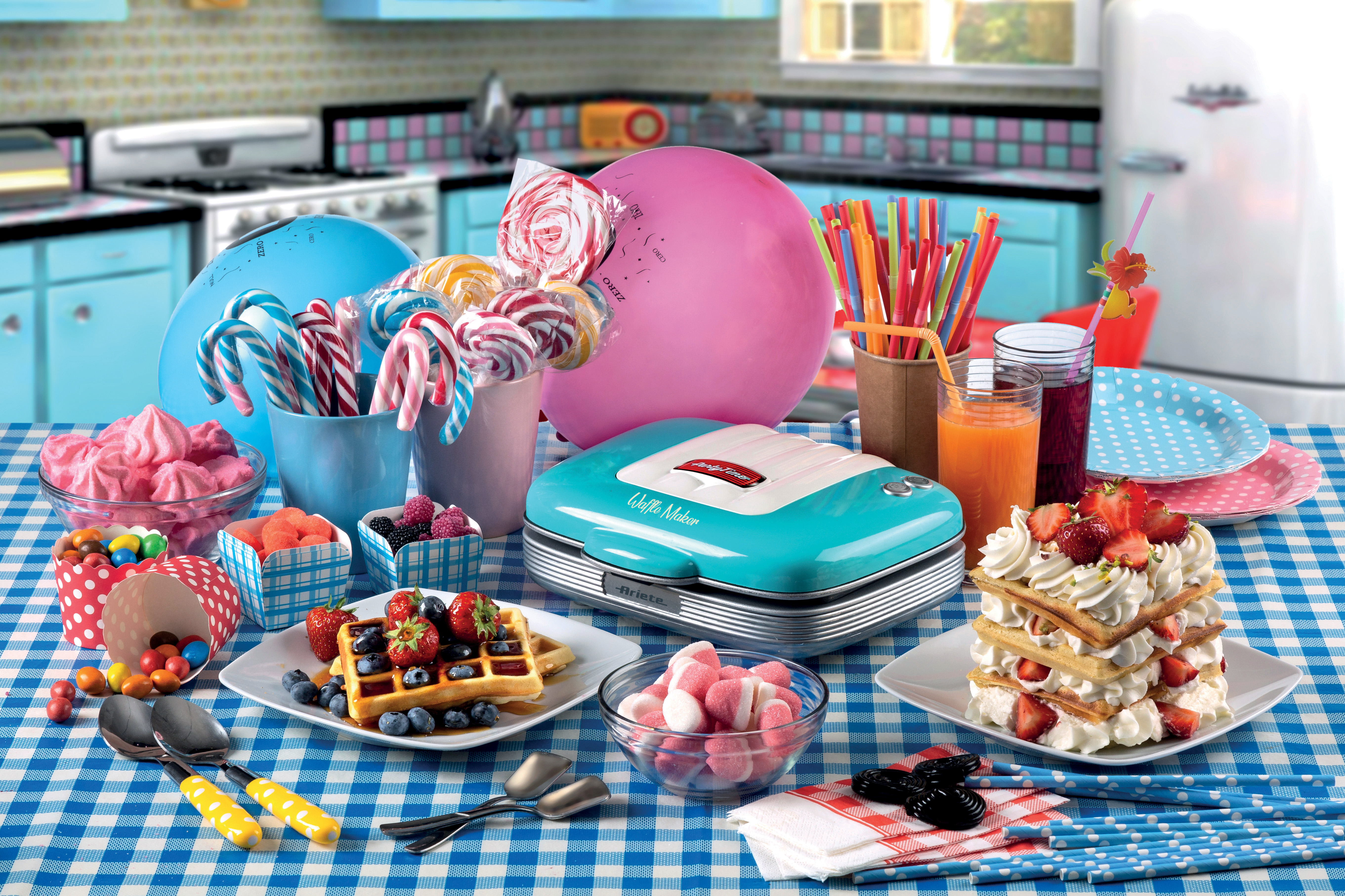 Una colorida mesa de fiesta tiene gofres, dulces, bebidas, globos y una gofrera turquesa.