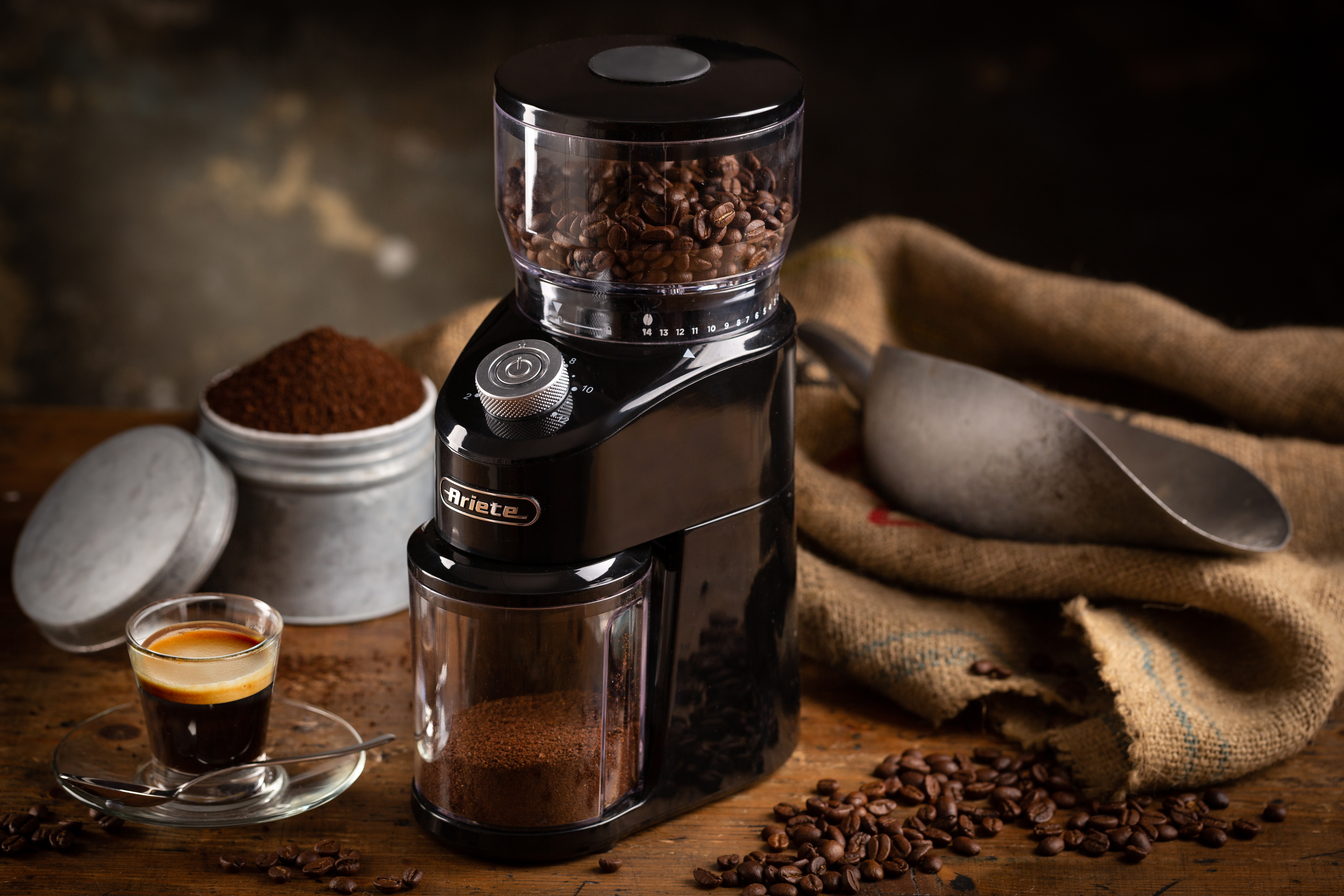 Molinillo de café negro en mesa de madera con granos, café molido en latas y una taza de espresso cerca.