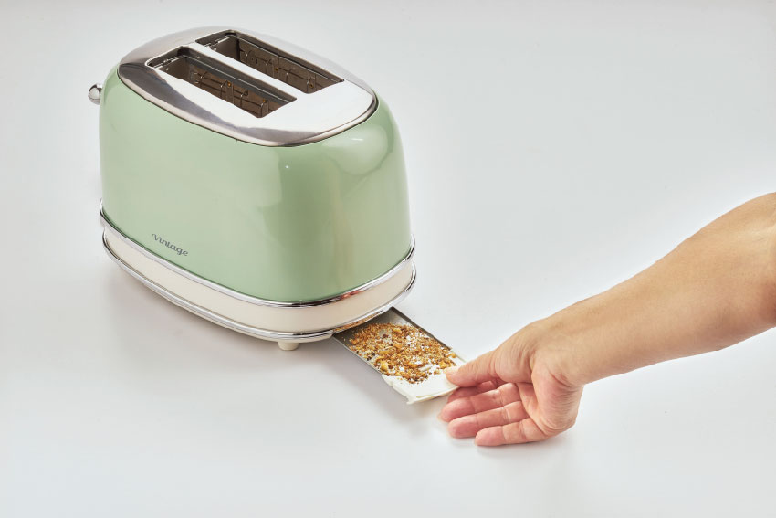 Eine Hand zieht die Krümelschublade eines grünen Vintage-Toasters auf weißer Fläche heraus.