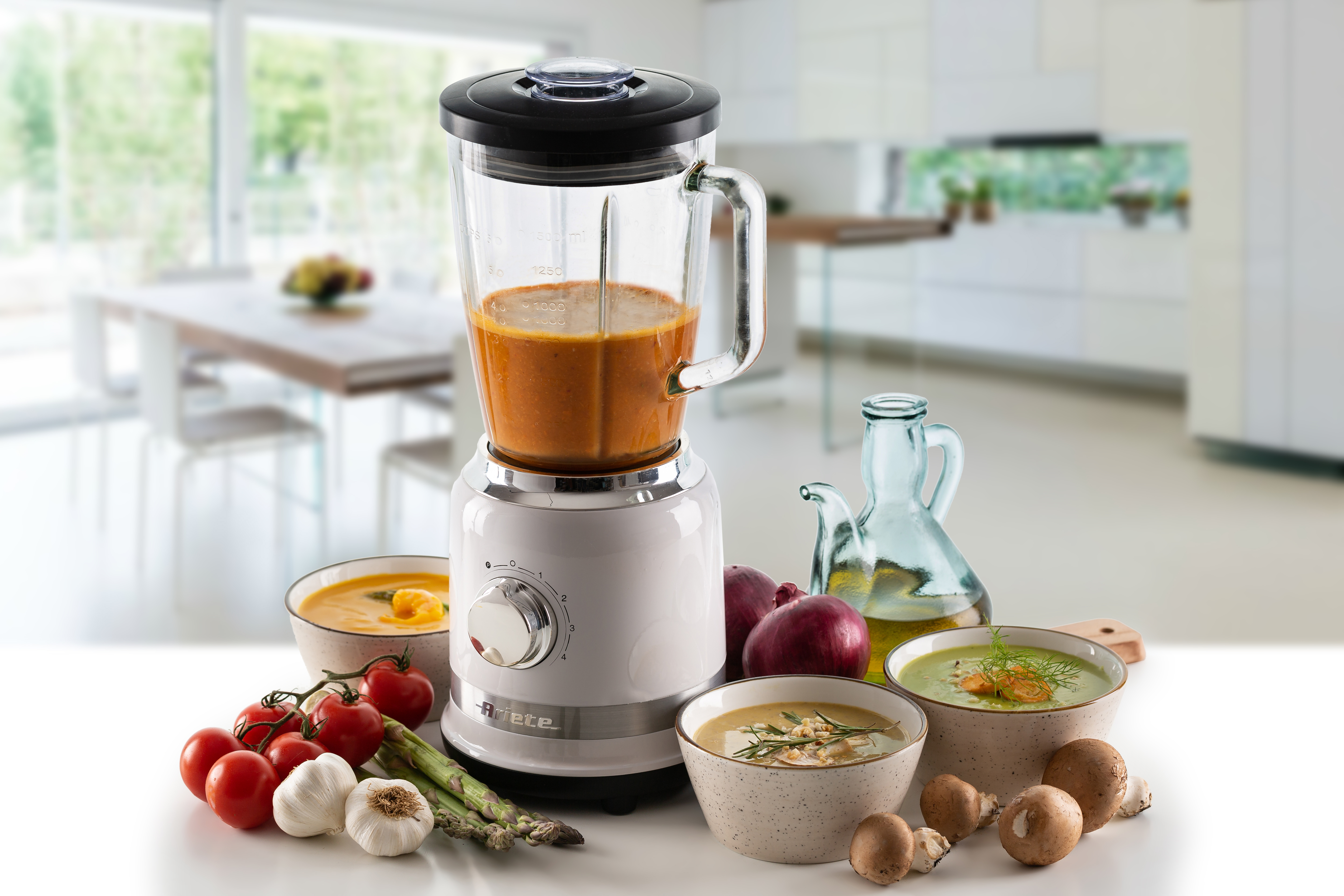 Bir mutfak tezgahında çorba dolu beyaz bir blender, taze sebzeler ve kaseler.