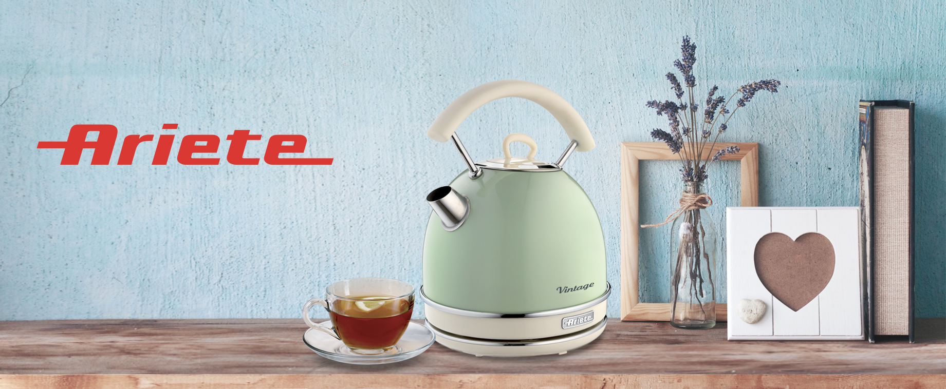 Ein pastellgrüner Vintage-Wasserkocher, eine Tasse Tee und Dekoartikel auf einem Holzregal vor einer blauen Wand.