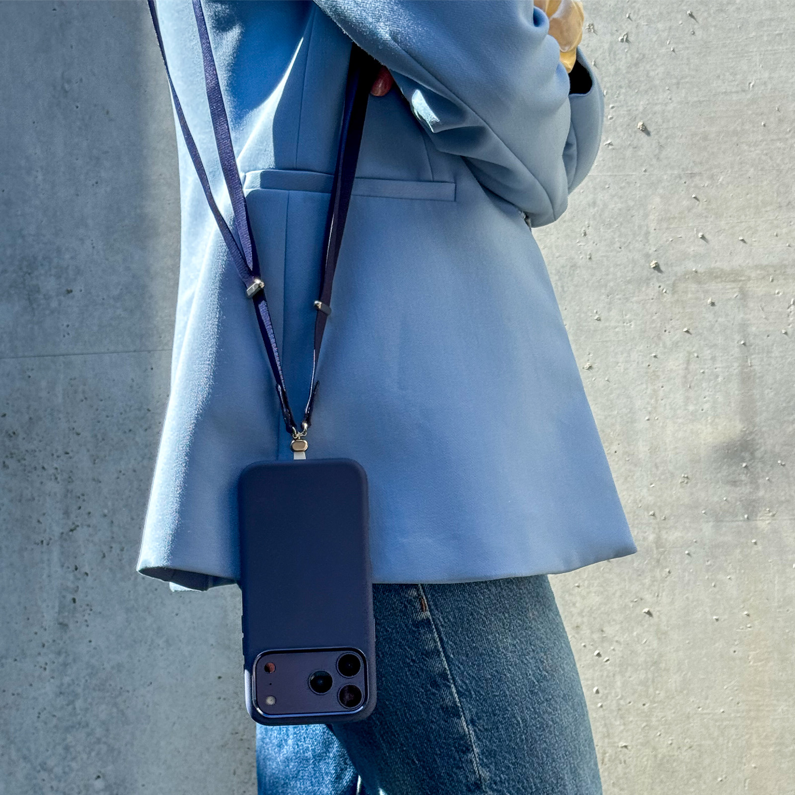 Una persona con blazer azul claro y vaqueros lleva un smartphone azul oscuro con correa ante una pared.