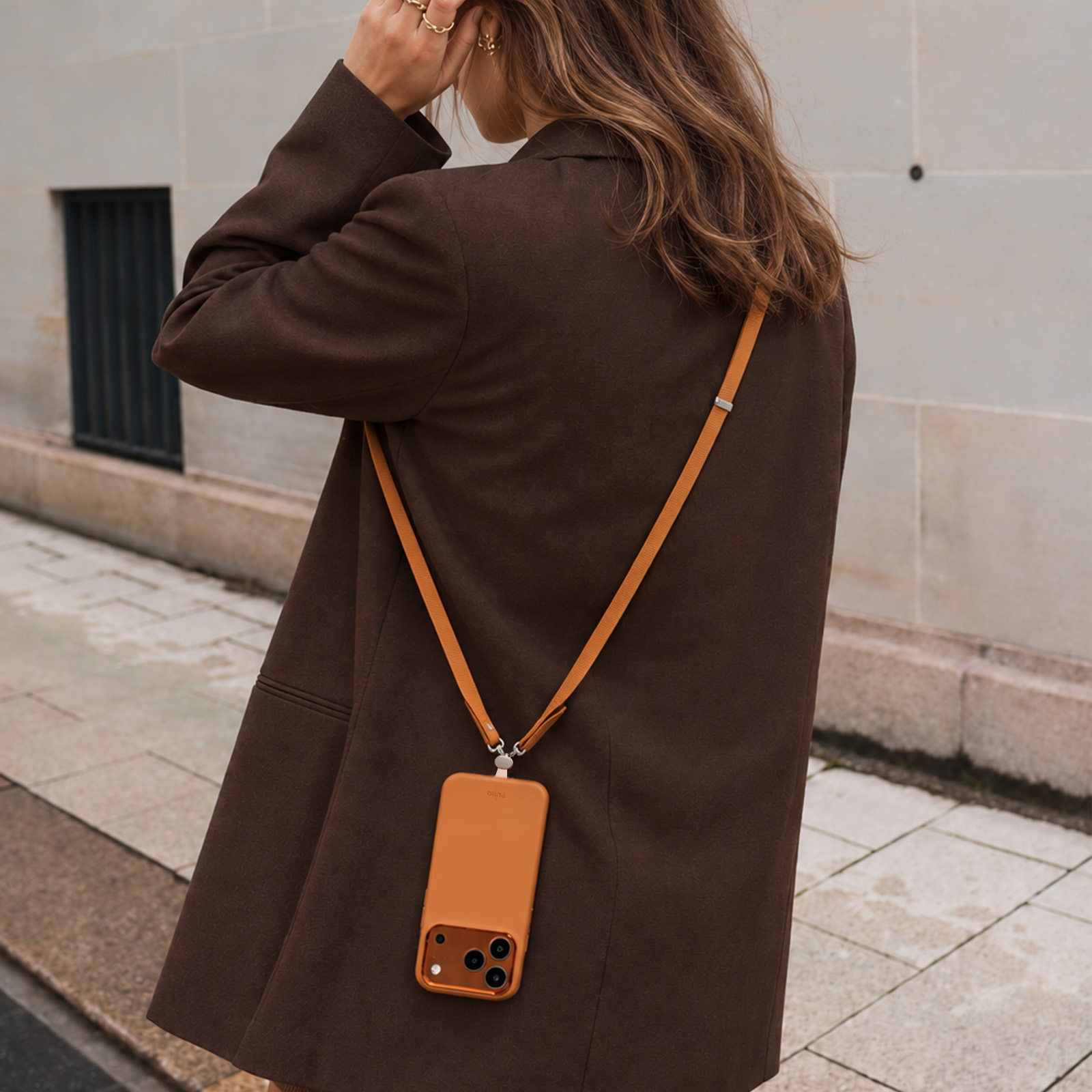Una mujer con blazer marrón lleva un smartphone marrón colgado del hombro al aire libre.