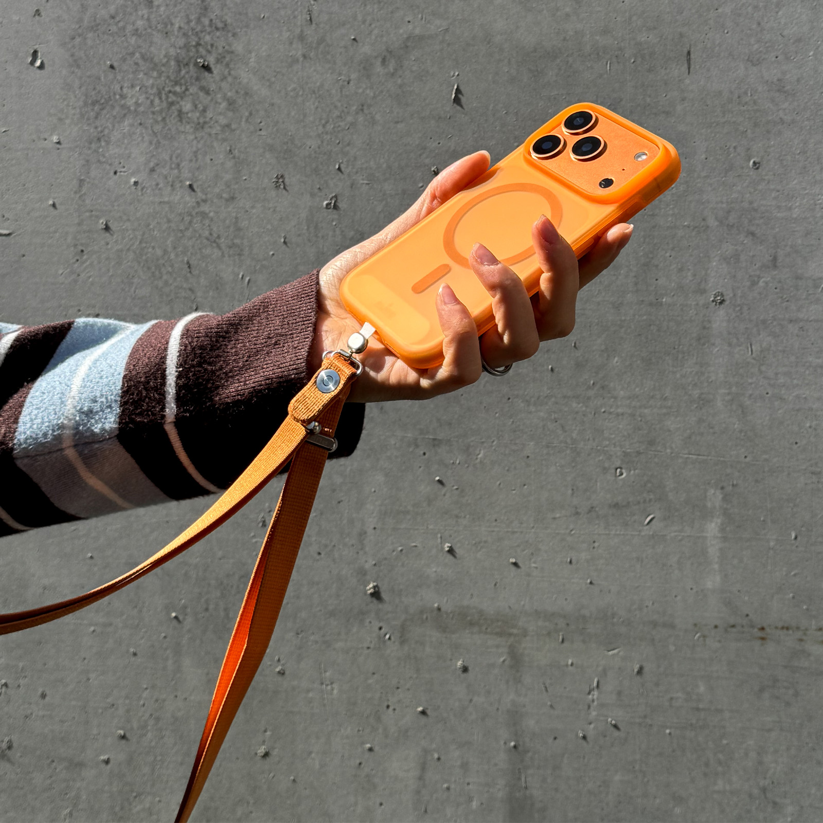 Una mano sostiene un smartphone naranja con funda y correa a juego frente a una pared de hormigón.