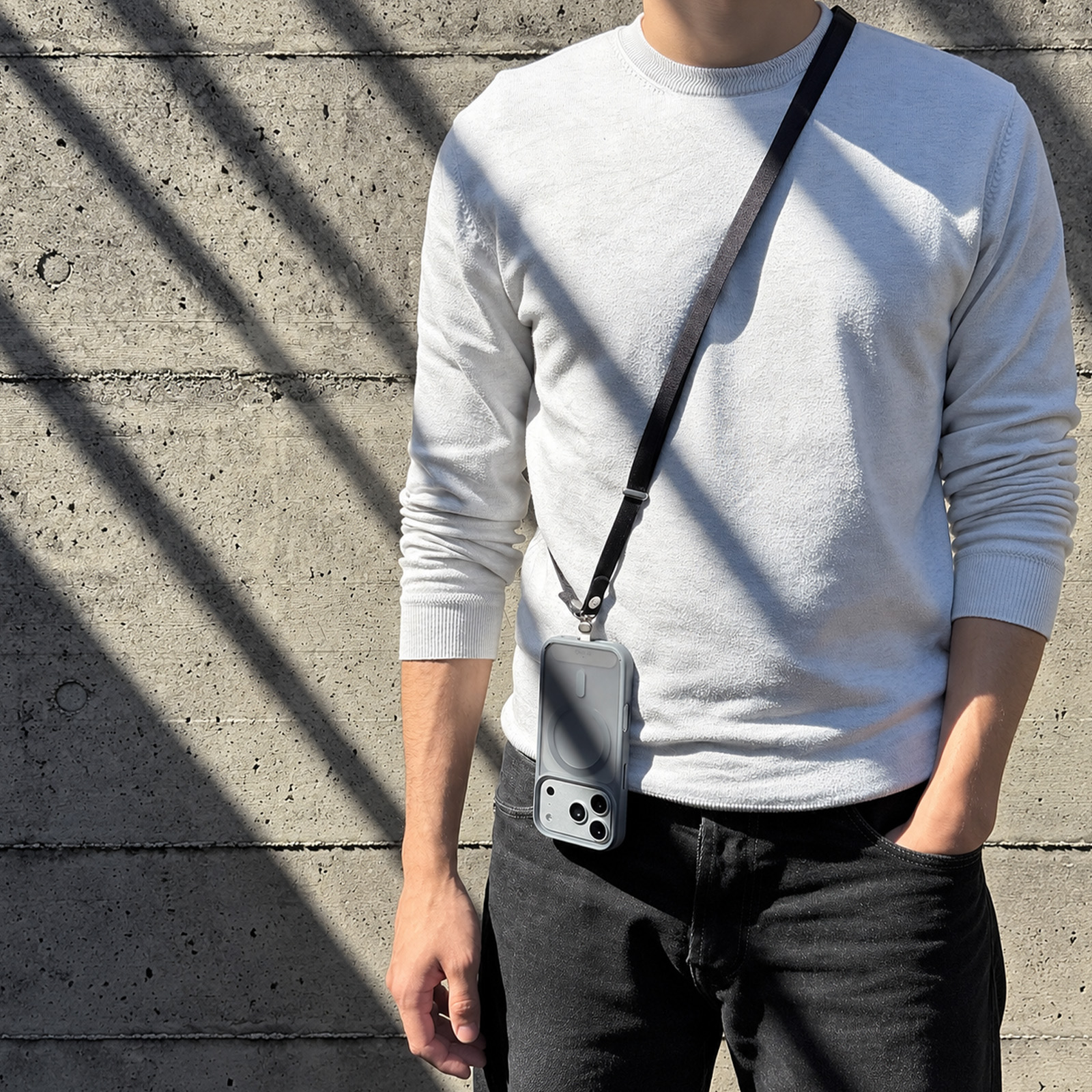 Un hombre con jersey blanco y vaqueros negros lleva un smartphone con cordón ante una pared de hormigón.