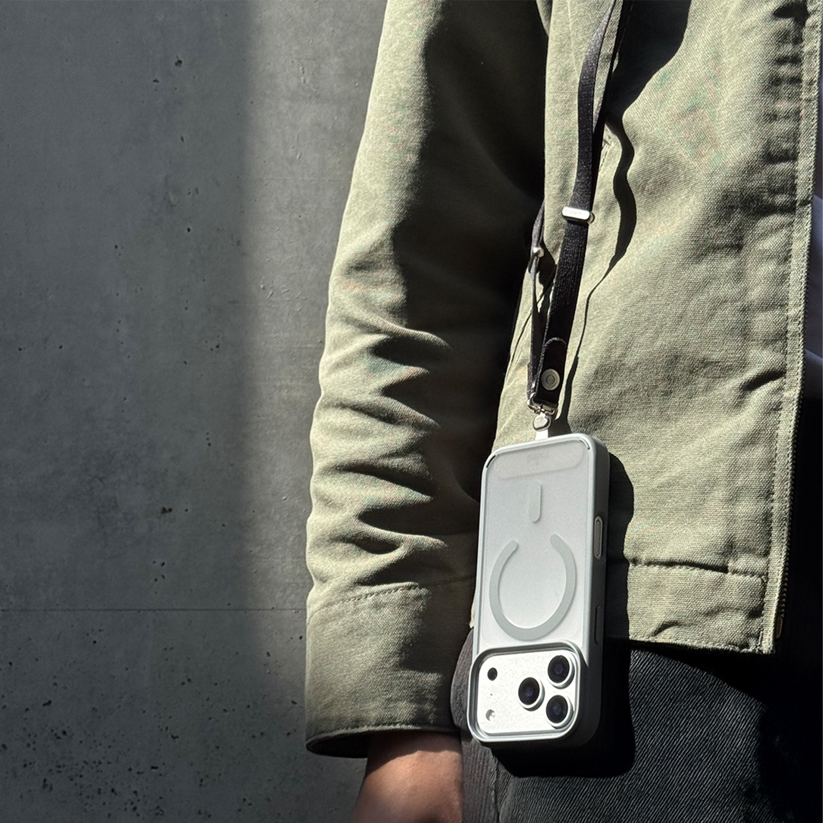 Una persona vestida de verde lleva un teléfono inteligente con correa ante una pared de hormigón gris.