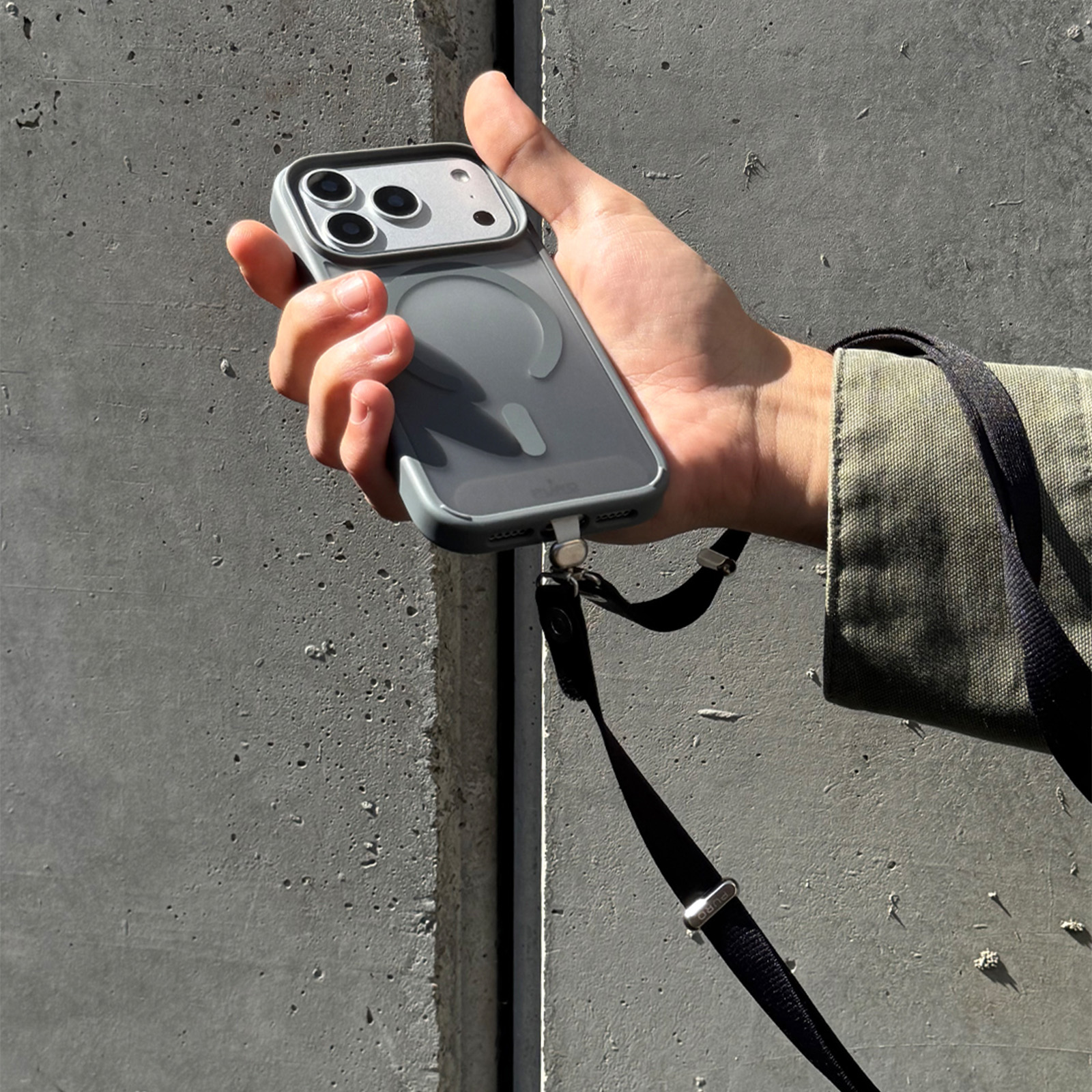 Mano sostiene un smartphone en funda gris con correa negra frente a una pared de hormigón.