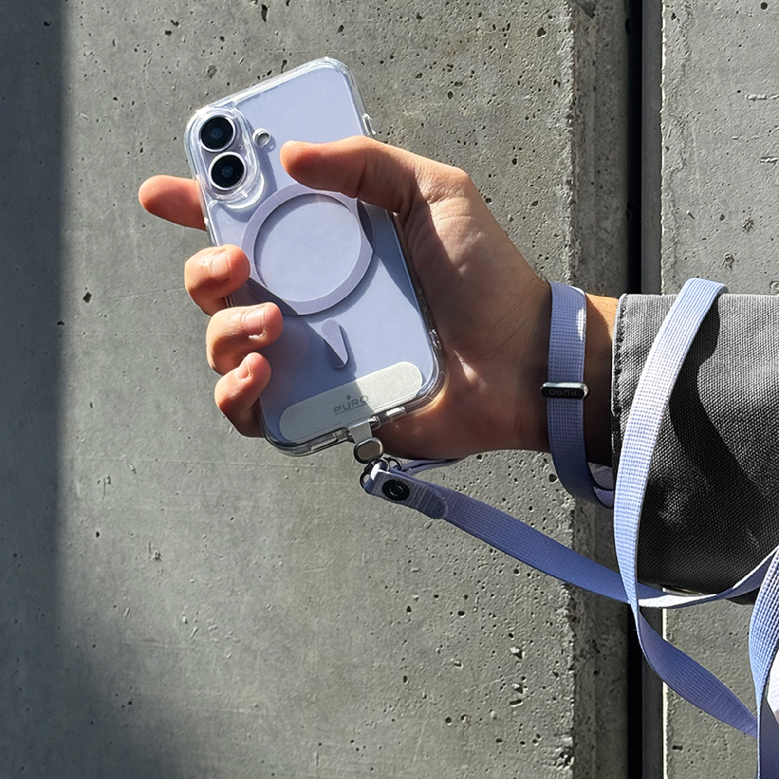 Una mano sostiene un teléfono celeste con funda MagSafe y correa frente a una pared de hormigón.