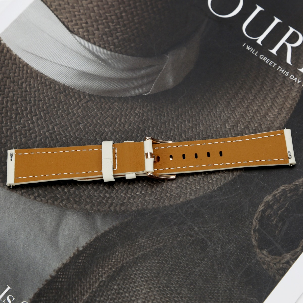 Ein braunes Lederarmband mit weißer Naht liegt waagerecht auf einer Magazinseite mit einem braunen Hut.