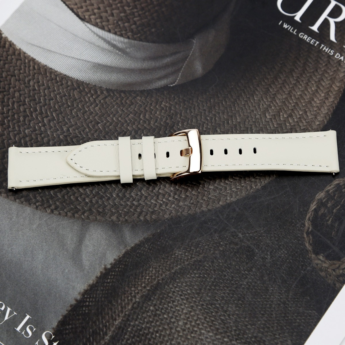 Ein weißes Lederarmband mit Roségold-Schließe auf strukturiertem dunklem Hintergrund.