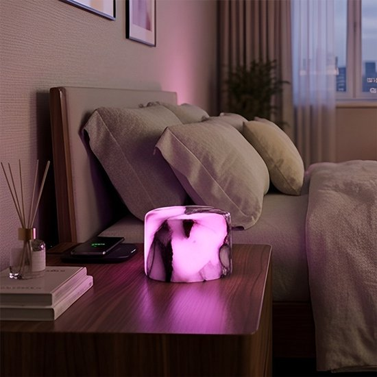 Eine leuchtend rosa zylindrische Lampe steht auf einem Nachttisch neben einem Bett mit Kissen.