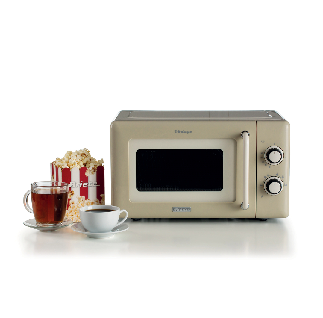 Un microondas beige de estilo retro sobre una mesa blanca junto a una caja de palomitas y dos tazas.