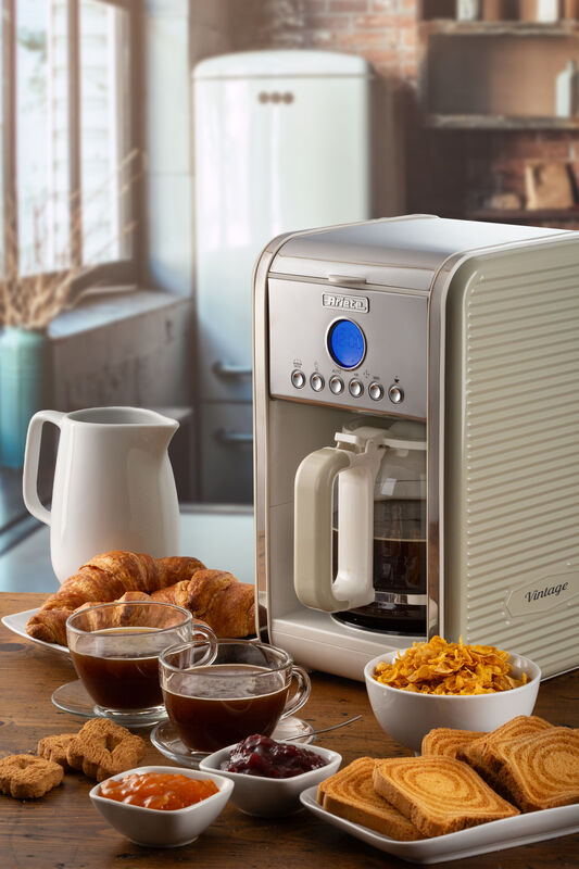 A beige coffee maker on a wooden table, surrounded by croissants, cups of coffee, jam, and toast slices.