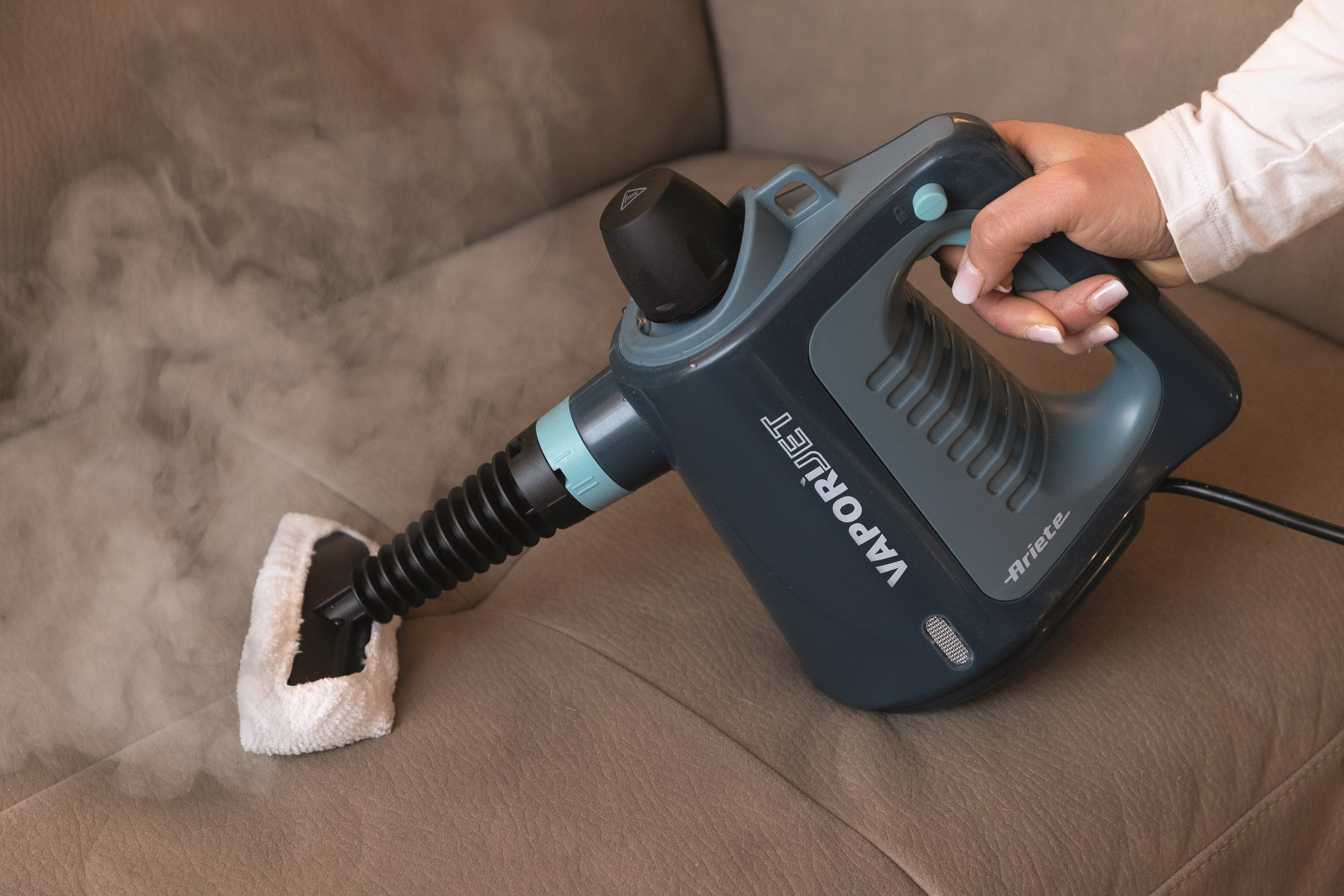 Eine Hand benutzt einen grauen Ariete Dampfreiniger, um ein braunes Sofa mit heißem Dampf zu reinigen.