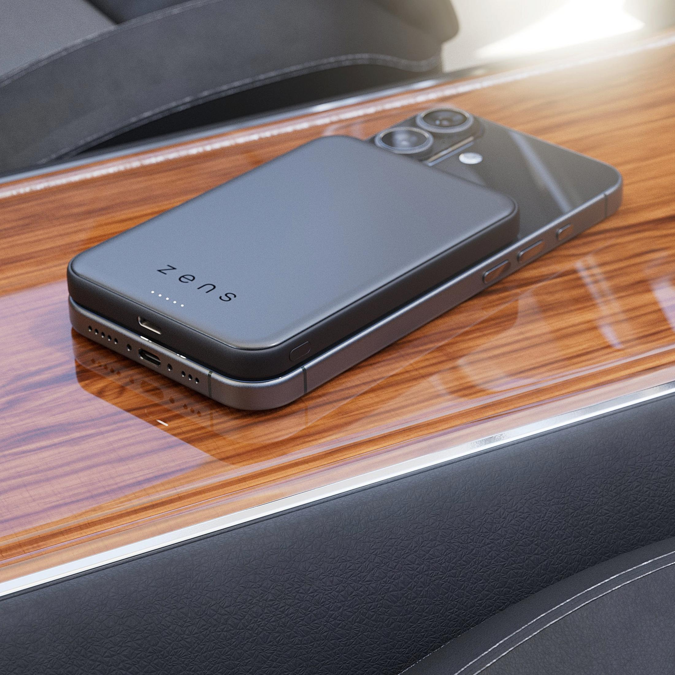 A black Zens power bank attached magnetically to the back of a smartphone resting on a wooden console.