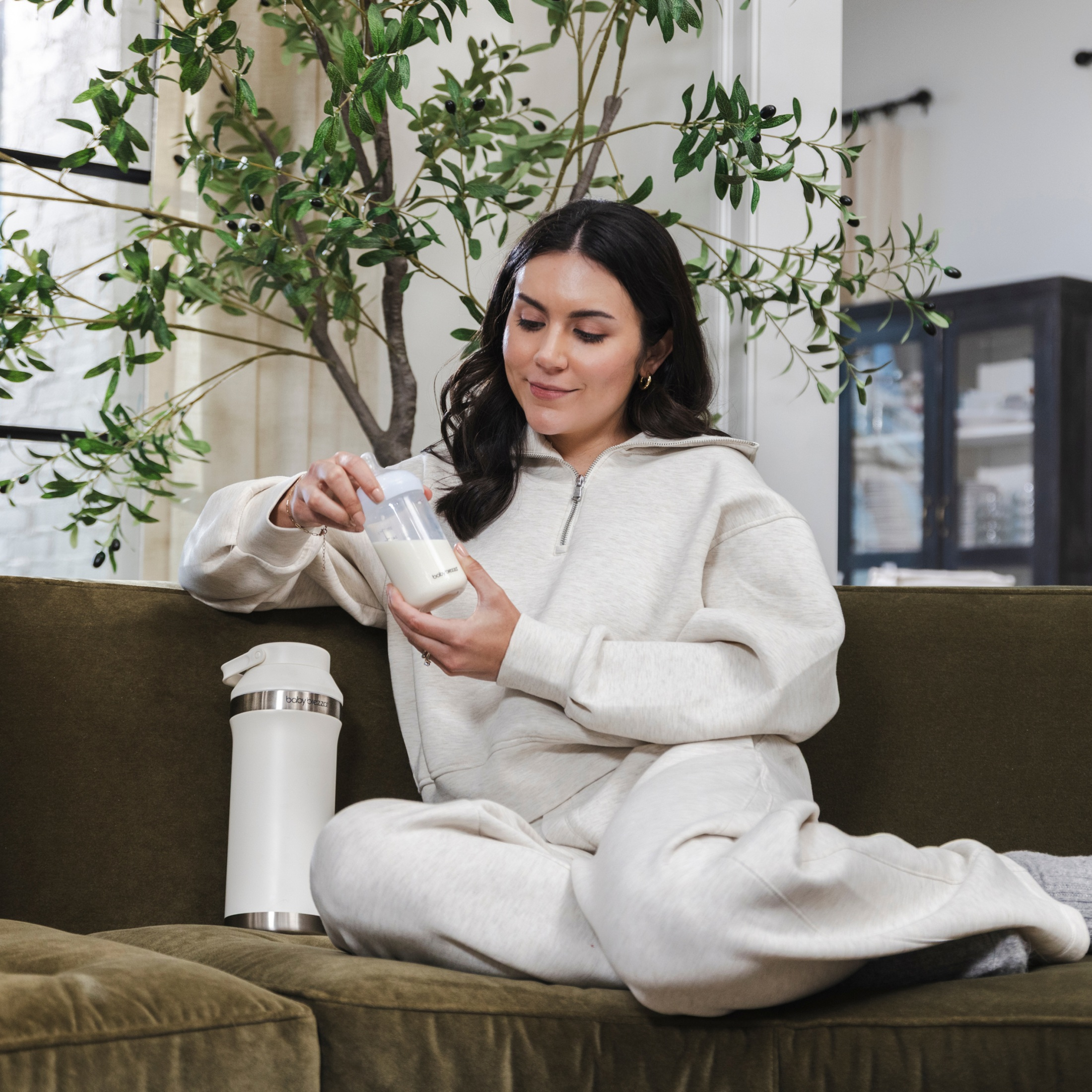 Eine Frau in weißer Loungewear sitzt auf einem grünen Sofa und hält eine Babyflasche vor einer Pflanze.