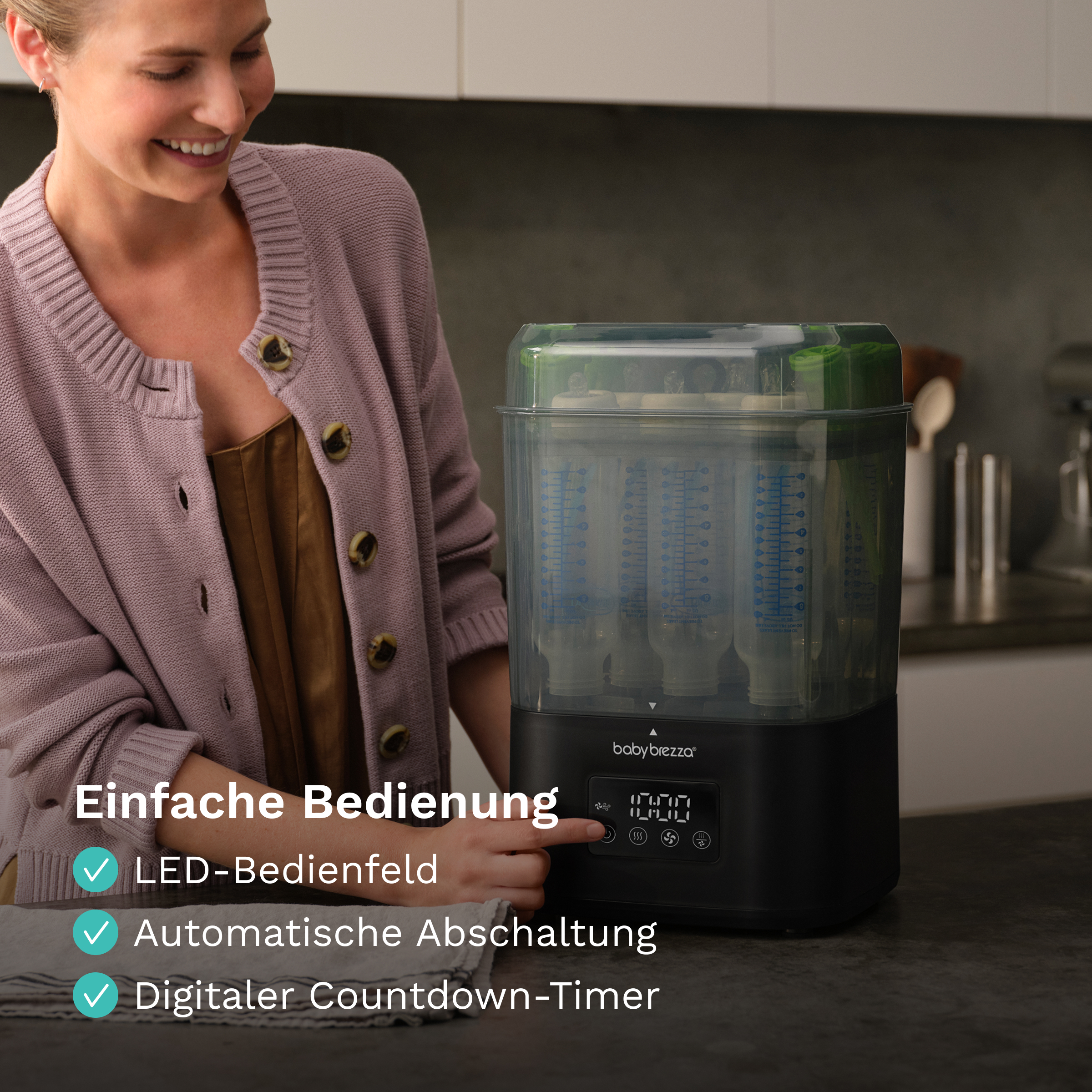 Eine Frau bedient einen schwarzen Babybrezza Flaschensterilisator in einer Küche mit Textoverlay.