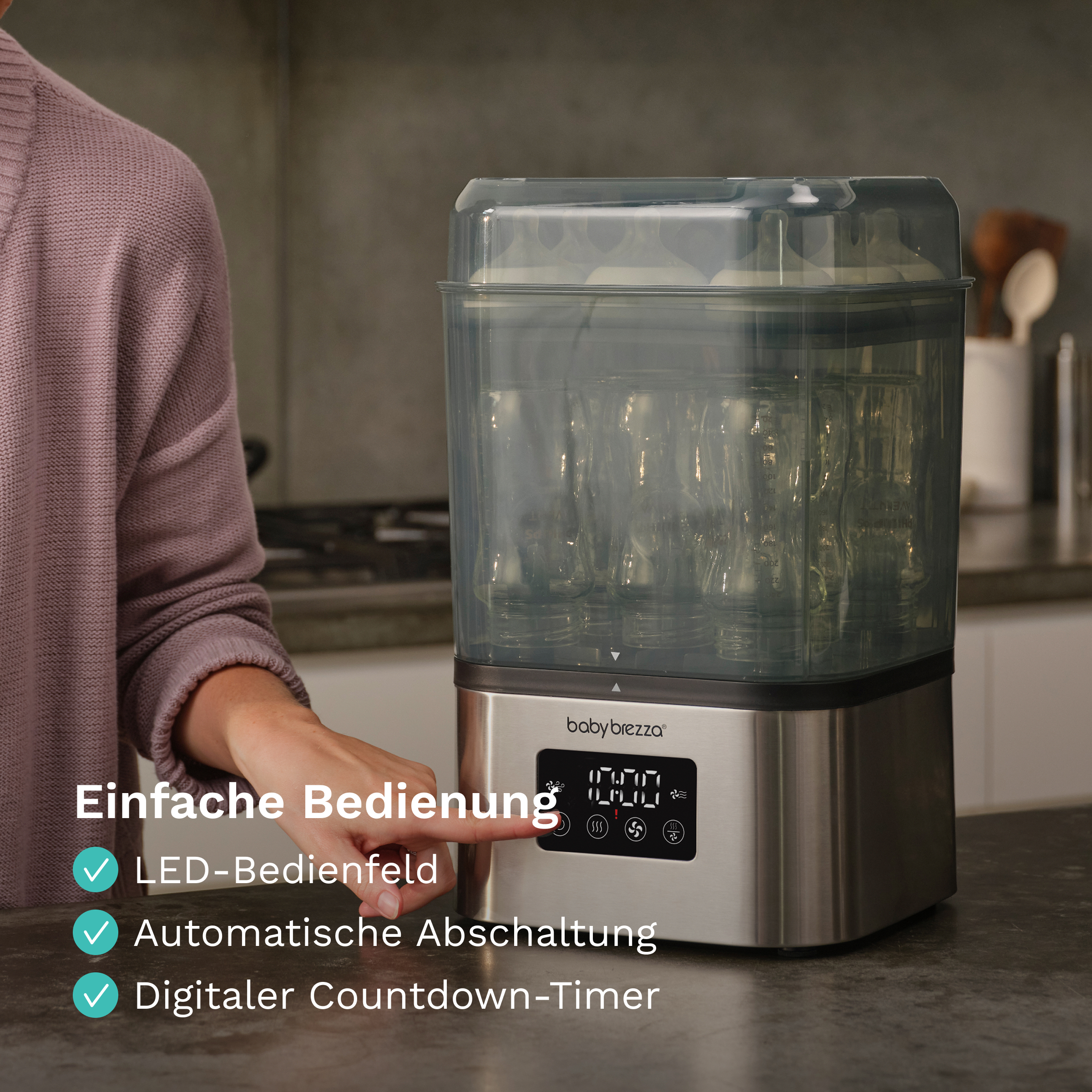 Eine Hand bedient das LED-Bedienfeld eines Baby Brezza Flaschensterilisators auf einer Küchentheke.