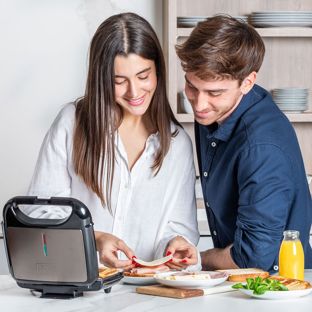 Paar lächelt beim Zubereiten von Sandwiches mit einem Sandwichmaker in der Küche.
