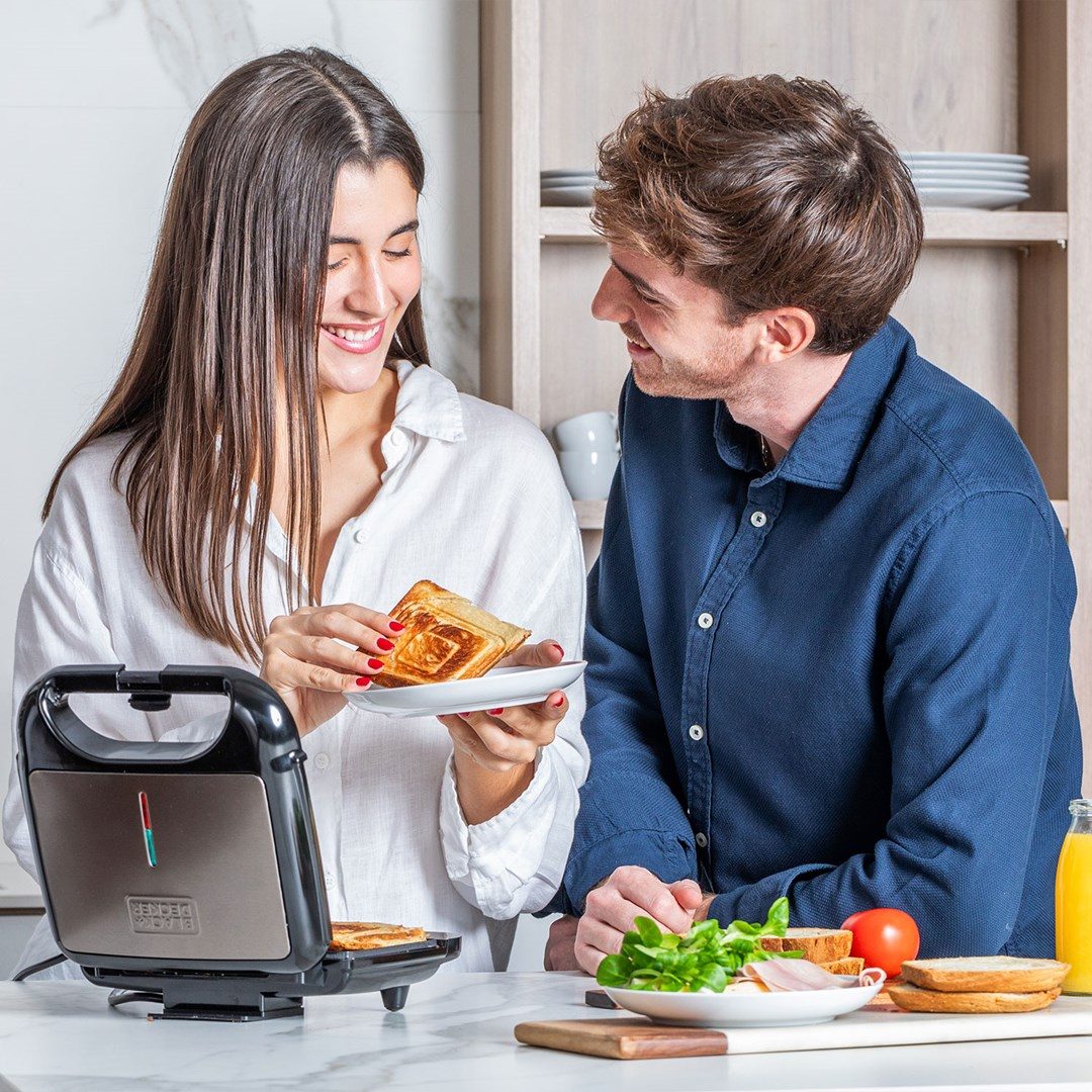 Ein glückliches Paar bereitet in einer hellen Küche Toasts mit einem Sandwichmaker zu.