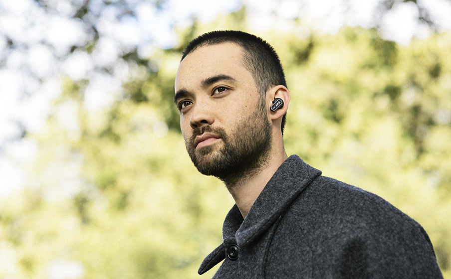 Ein junger Mann mit Bart trägt schwarze In-Ear-Kopfhörer vor einer grünlichen Naturkulisse.