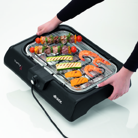 Hands holding an electric tabletop grill with skewers, shrimp, and vegetables.