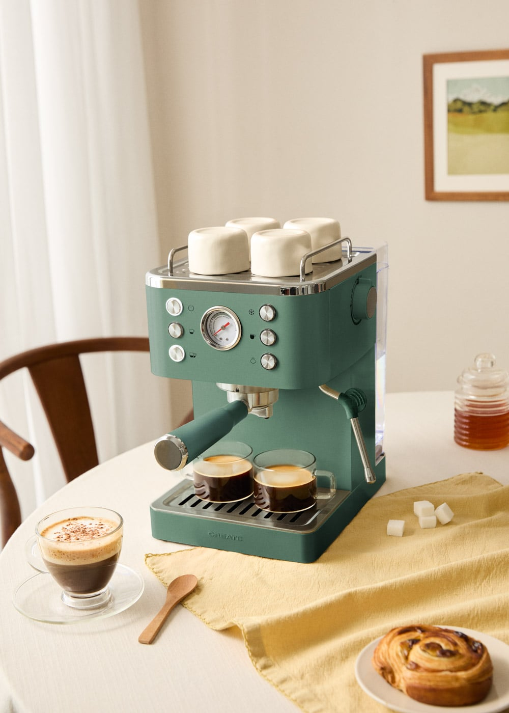 Una cafetera espresso verde en una mesa con dos tazas de café, terrones de azúcar y repostería cerca.
