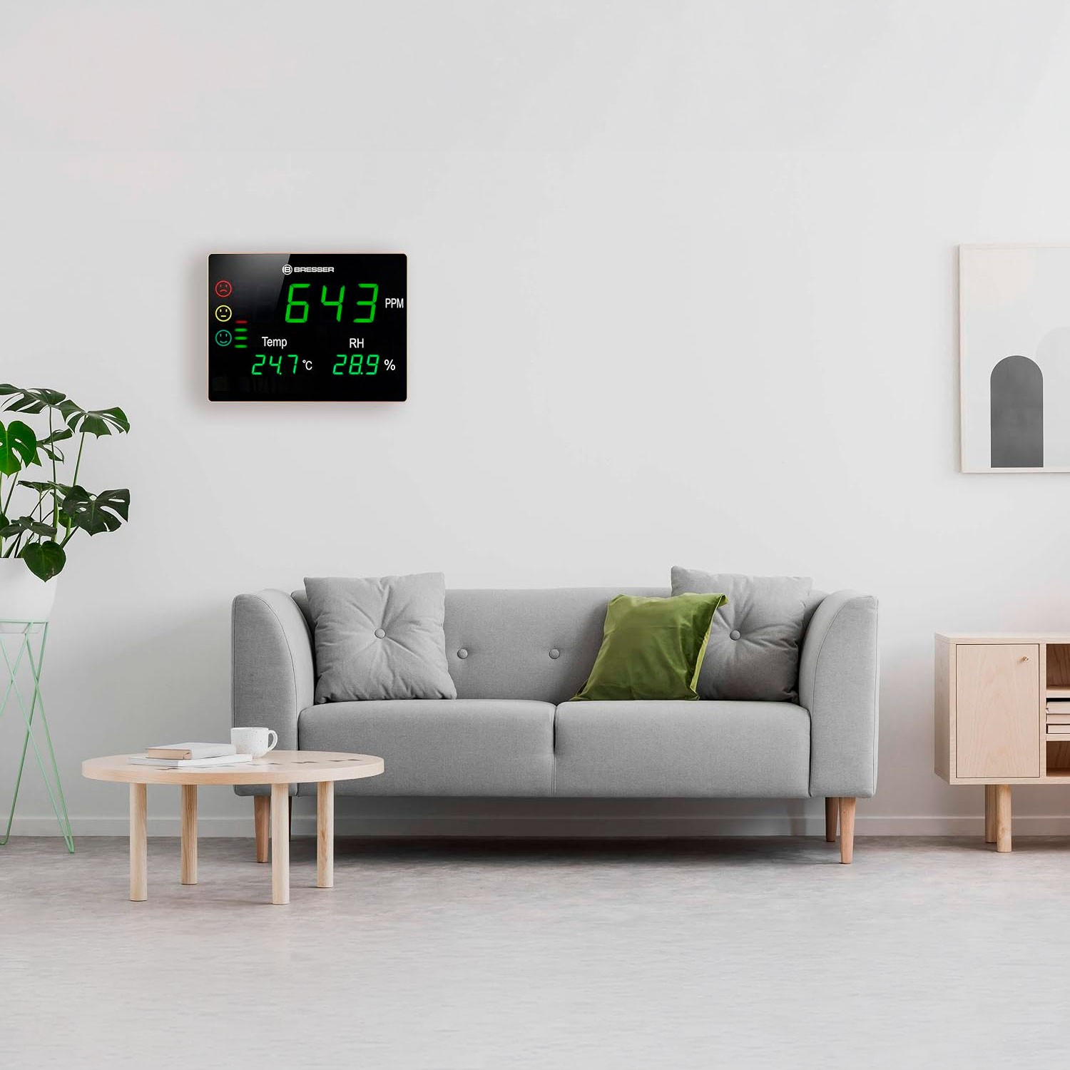 Un sofá gris, mesa de madera, planta y un medidor digital de CO2 en una pared blanca de una sala moderna.